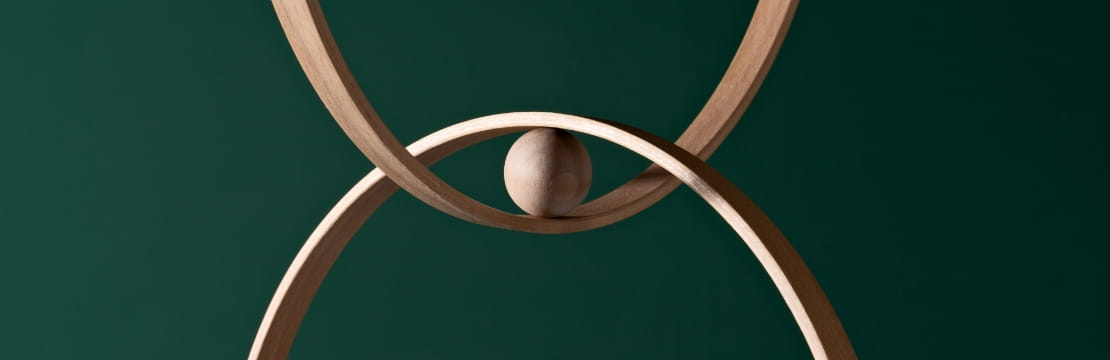Two intersecting wooden rings with a small wooden sphere balanced at their meeting point, set against a dark green background.